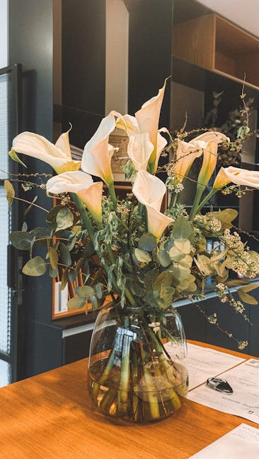 A large, elegant floral arrangement featuring creamy white calla lilies with smooth, curved petals and prominent yellow spadices, complemented by soft green eucalyptus leaves and small white filler fl