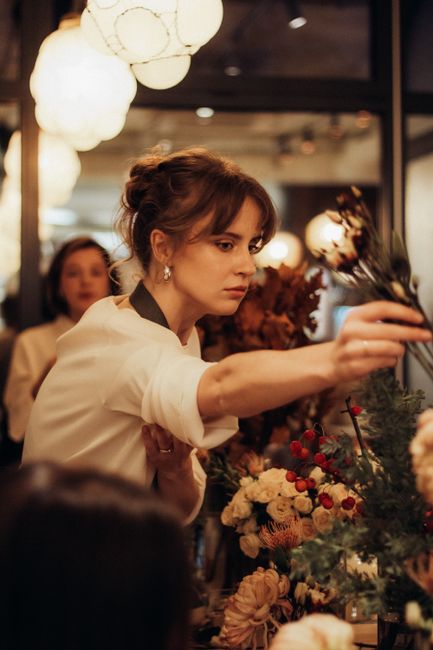 A woman with brown hair styled in a loose updo, wearing a cream-colored top, is carefully arranging a floral display or bouquet at a flower shop or event setting. She is extending her arm to position