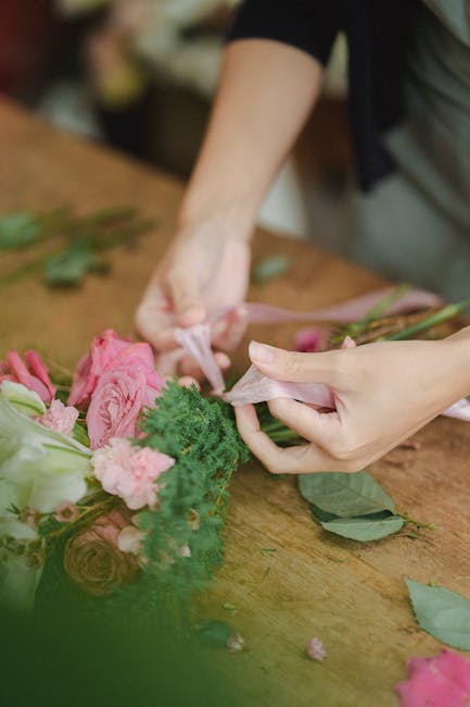 A close-up of a florist's hands arranging a bouquet of fresh flowers on a wooden table. The bouquet includes pink roses, green foliage, and delicate pink carnations, all with vibrant, healthy petals a